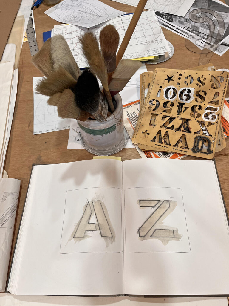 Image from the studio during the creation of the Stephen Zaima painting "AZ" (2023) depicting a sketchbook with lettering for the painting, letter stencils and paintbrushes.